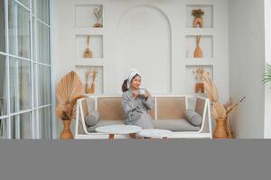 Happy attractive Asian woman in grey bathrobe posing at the terrace, smiling at camera while drinking tea. Holiday leisure concept. photo