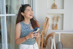 Portrait of a cheerful young Asian woman standing at terrace, smiling and holding smartphone while wearing casual outfit on holiday photo