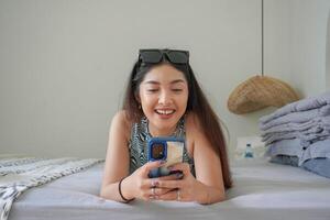 sonriente asiático mujer tendido abajo en cama con Gafas de sol en parte superior mientras utilizando móvil teléfono, explorador social medios de comunicación en el Mañana foto