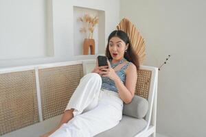 conmocionado asiático mujer participación y viendo teléfono con boca amplio abierto mientras tendido en un sofá en un villa para fiesta foto