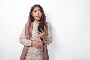 Young Asian Muslim woman in veil hijab feeling hungry while holding her stomach and using a smartphone to order foods. Ramadan and Eid Mubarak concept photo