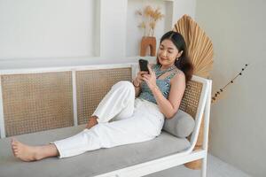 retrato de contento asiático mujer sonriente y participación su teléfono inteligente mientras tendido abajo en un sofá en villa para verano fiesta foto