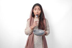 Hungry Asian Muslim woman holding bowl thinking what to eat with copy space over the bowl while biting spoon waiting for iftar, isolated white background photo