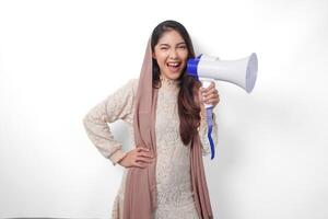 animado joven asiático musulmán mujer vistiendo Pañuelo velo hijab gritos a megáfono, aislado en blanco antecedentes estudio. Ramadán y eid Mubarak concepto. foto