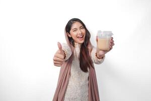 Excited Asian Muslim woman in headscarf veil hijab giving thumbs up while holding coffee in a plastic cup waiting for iftar to drink. Ramadan concept photo