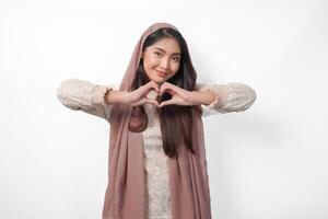 A happy young Asian Muslim woman wearing veil hijab making love heart shape using hands on isolated white background. Ramadan and Eid Mubarak concept photo