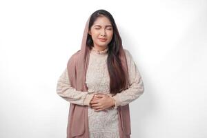 Portrait of Asian Muslim woman wearing headscarf veil placing hands on the stomach feeling hungry and want to eat while fasting, isolated over white background. Ramadan and Eid Mubarak concept photo