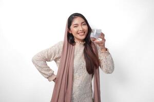 Young Asian Muslim woman wearing headscarf veil smiling to the camera and holding glass with water, isolated by white background. Ramadan concept photo