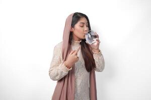 joven asiático musulmán mujer vistiendo Pañuelo velo sensación sediento y Bebiendo agua después ayuno, aislado por blanco antecedentes. Ramadán concepto foto