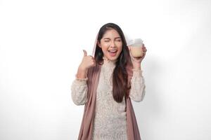 Excited Asian Muslim woman in headscarf veil hijab giving thumbs up while holding coffee in a plastic cup waiting for iftar to drink. Ramadan concept photo