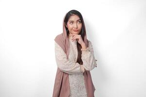 Thoughtful Asian Muslim woman wearing headscarf veil hijab focused and looking to copy space beside her, standing on isolated white background photo