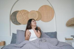 Happy attractive Asian woman in white top posing at the bedroom after waking up, smiling cheerfully while holding cup of tea. Holiday leisure concept. photo