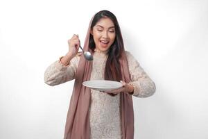 Portrait of hungry Asian Muslim woman holding cutlery and plate with copy space, feeling excited to eat after fasting on isolated white background. Ramadan concept photo