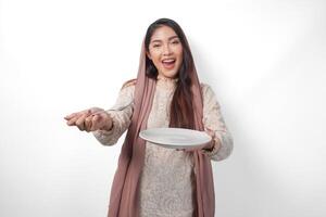 Excited Asian Muslim woman holding plate and cutlery to eat after fasting while smiling cheerfully on isolated white background. Ramadan concept photo