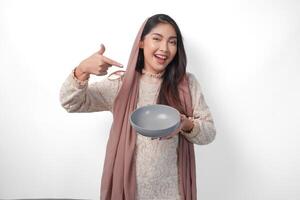 Portrait of beautiful Asian Muslim woman in headscarf veil hijab smiling wide and pointing to the bowl she holds with copy space inside. Ramadan concept photo