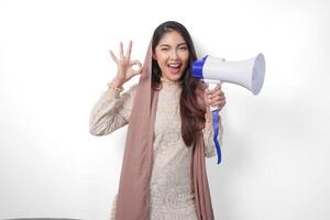 Attractive young Asian Muslim woman in veil hijab gesturing OK okay, good sign with her fingers while shouting at megaphone. Ramadan and Eid Mubarak concept photo