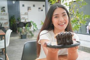 joven asiático mujer participación y demostración un negro plato de rosquilla Pastelería llamado cromboloni con chocolate relleno para bocadillo tiempo, sonriente con un contento y alegre expresión foto