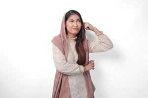 Thoughtful Asian Muslim woman wearing headscarf veil hijab focused and looking to copy space beside her, standing on isolated white background photo