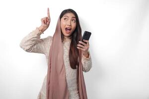 Surprised Asian Muslim woman in veil hijab holding a smart phone while pointing up to the copy space upwards over isolated white background. Ramadan concept photo