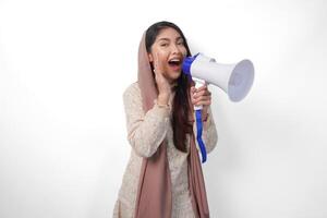 Lively Young Asian muslim woman wearing headscarf veil hijab shouting at megaphone, isolated on white background studio. Ramadan and Eid Mubarak concept. photo