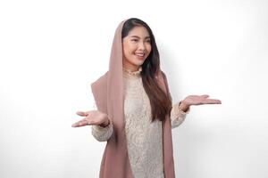 Smiling Asian Muslim woman in headscarf veil hijab presenting copy space, standing over isolated white background. Ramadan and Eid Mubarak advertisement concept photo