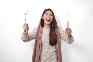 Portrait of Asian Muslim woman in veil hijab holding cooking tools or utensils while smiling cheerfully over isolated white background. Ramadan and Eid Mubarak concept photo