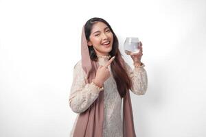 Portrait of beautiful Asian Muslim woman in headscarf veil hijab smiling wide and pointing to the glass of water she holds. Ramadan concept photo