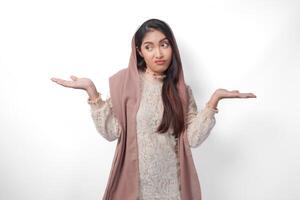 Confused Asian Muslim woman wearing headscarf veil hijab thinking and contemplating choices while pointing to copy space beside her, standing on isolated white background photo