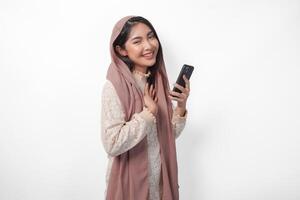 Happy mindful thankful young Asian Muslim woman hand on chest smiling while holding smartphone, isolated on white background photo