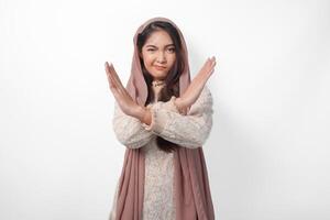 Young Asian Muslim woman wearing veil hijab gesturing a no rejection, refuse using hands and fingers, isolated by white background. Ramadan and Eid Mubarak concept photo