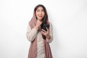 hambriento asiático musulmán mujer en velo hijab pensando qué a comer después rápido mientras utilizando un teléfono inteligente a orden alimentos Ramadán concepto foto