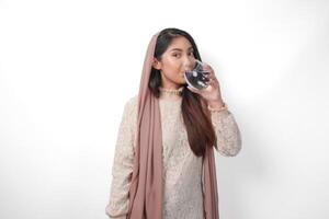 Young Asian Muslim woman wearing headscarf veil feeling thirsty and drinking water after fasting, isolated by white background. Ramadan concept photo