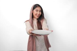 Excited Asian Muslim woman holding plate and cutlery to eat after fasting while smiling cheerfully on isolated white background. Ramadan concept photo