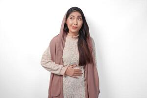 Portrait of Asian Muslim woman wearing headscarf veil placing hands on the stomach feeling hungry and want to eat while fasting, isolated over white background. Ramadan and Eid Mubarak concept photo