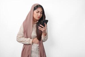 joven asiático musulmán mujer en velo hijab sensación hambriento mientras participación su estómago y utilizando un teléfono inteligente a orden alimentos Ramadán y eid Mubarak concepto foto