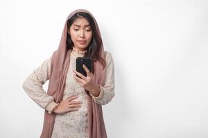joven asiático musulmán mujer en velo hijab sensación hambriento mientras participación su estómago y utilizando un teléfono inteligente a orden alimentos Ramadán y eid Mubarak concepto foto