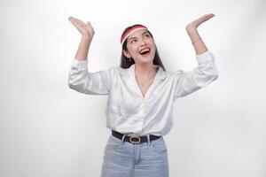 Happy Asian woman wearing Indonesia flag colored headband and casual outfit while smiling cheerfully and pointing up the copy space upwards. Independence day advertisement concept photo