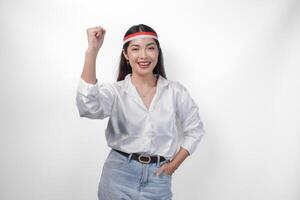 Excited young Asian woman wearing country flag headband and clenching hands while celebrating Indonesian independence day isolated on white background photo