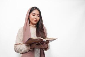 Serious Asian Muslim woman wearing veil hijab praying and reading Al Quran, standing over isolated white background. Ramadan and Eid Mubarak concept photo