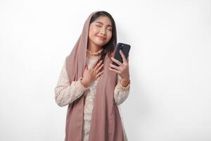 contento consciente de agradecido joven asiático musulmán mujer mano en cofre sonriente mientras participación teléfono inteligente, aislado en blanco antecedentes foto