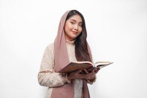 Serious Asian Muslim woman wearing veil hijab praying and reading Al Quran, standing over isolated white background. Ramadan and Eid Mubarak concept photo