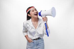 Energetic Asian woman in casual formal outfit wearing country flag headband while holding and shouting at megaphone, standing on isolated white background photo