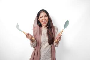 Portrait of a happy Asian Muslim woman holding kitchen cooking utensils while smiling cheerfully on isolated white background. Ramadan concept photo