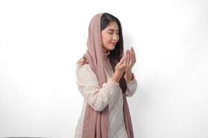 Religious Asian Muslim woman wearing veil headscarf with hands in praying gesture to God, standing over isolated white background. Ramadan and Eid Al Fitr concept photo