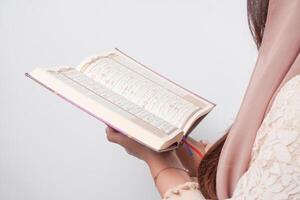 Close up portrait of Asian Muslim woman in headscarf veil hijab holding and reading Al Quran over isolated white background. Ramadan and Eid Mubarak concept. photo