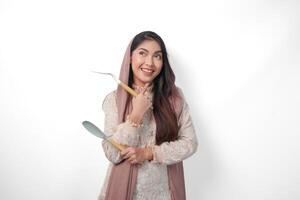 Thoughtful Asian Muslim woman wearing headscarf veil hijab focused and looking to copy space beside her while holding spatula or kitchen utensils, standing on isolated white background photo