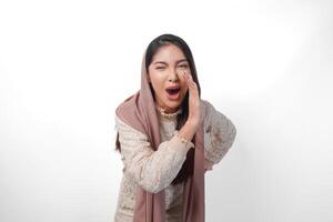 joven asiático mujer vistiendo Pañuelo velo hijab es levantamiento manos y gritos con boca amplio abierto, aislado blanco antecedentes foto