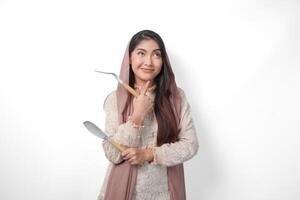 Thoughtful Asian Muslim woman wearing headscarf veil hijab focused and looking to copy space beside her while holding spatula or kitchen utensils, standing on isolated white background photo