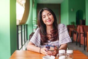 sonriente joven asiático mujer vistiendo vestir y sensación feliz, sentado a un restaurante para un desayuno mientras participación un teléfono inteligente foto