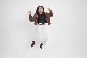 activo asiático niña es saltando y levantamiento manos arriba vestir marrón chaqueta, aislado blanco color antecedentes foto